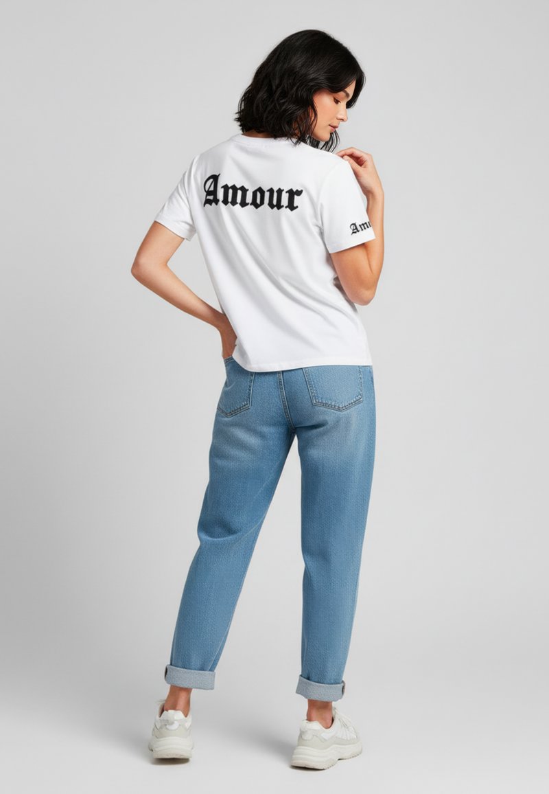 Witte katoenen t-shirt met zwarte tekst "Amour" op de achterkant en mouw, gecombineerd met lichtblauwe denim jeans met opgerolde mouwen en witte sneakers.
