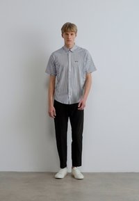 Cămașă cu mânecă scurtă, cu dungi, cu guler cu nasturi, asortată cu pantaloni negri conici și adidași albi, stând în fața unui perete simplu.