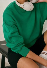 Groene sweatshirt met ribgebreide manchetten en schoudernaden, gedragen met beige hoofdtelefoon; gecombineerd met zwarte shorts en wit gestreepte sokken.