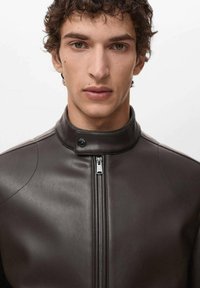 Veste en cuir marron avec un col montant, fermeture éclair et surface texturée. Présente un design épuré avec des accents matériels minimaux.
