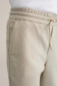 Gai bēšas jogas bikses, izgatavotas no faktūru auduma, ar elastīgu jostu un auklu, kā arī sānu kabatām.