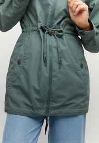 Zielona kurtka z wcięciem w talii, zapinana na zamek z przodu, z dwoma bocznymi kieszeniami. Posiada srebrne elementy ozdobne oraz gładkie, teksturowane wykończenie materiału.