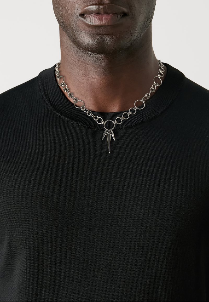 Collana in argento con maglie circolari e accenti a forma di punta, indossata sopra un maglione nero a collo rotondo.