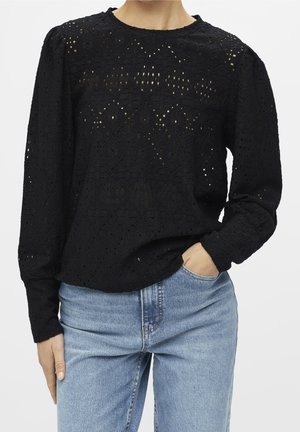 Blouse noire en dentelle à manches longues avec des motifs floraux, associée à un jean taille haute bleu clair, une main dans la poche, sur un fond uni.