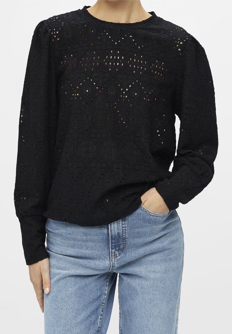Blouse noire en dentelle à manches longues avec des motifs floraux, associée à un jean taille haute bleu clair, une main dans la poche, sur un fond uni.