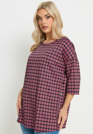 Donna con lunghi capelli biondi che indossa una tunica a quadri rosa e nera con maniche a tre quarti e dettagli con bottoni laterali.