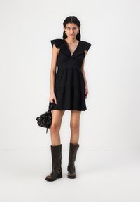 Vestido preto de algodão com mangas com folhos e um decote V profundo; saia em camadas; acompanhado de botas castanhas até ao joelho e uma mala preta com padrão.