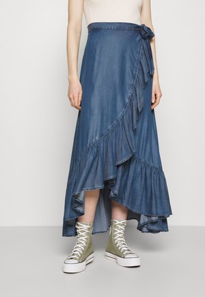 Spódnica kopertowa z denimu w ciemnym kolorze niebieskim z falbaną na dole i wiązaniem w talii, zestawiona z zielonymi sneakersami na wysokiej podeszwie.