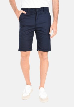 Pantaloni corti blu navy con design frontale piatto, orlo risvoltato e tasche laterali. Realizzati in un tessuto leggero, abbinati a scarpe da ginnastica bianche.