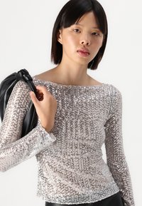 Μακρυμάνικη τοπ με παγιέτες σε ασημί αποχρώσεις, που διαθέτει στρογγυλή λαιμόκοψη και διάφανο ύφασμα. Ένα μοντέλο κρατά μια μαύρη δερμάτινη τσάντα στον ώμο.