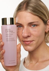 Estee Lauder Soft Clean Infusion-flaska, rosa med ett guldlock, hålls av en person. Texten säger "Hydrating Essence Lotion" och detaljer på engelska och franska.