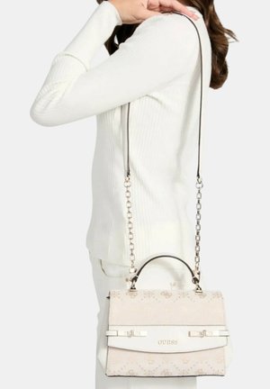 Donna che indossa un maglione a coste bianco e pantaloni che porta una borsa beige Guess con tracolla a catena argentata sulla spalla.