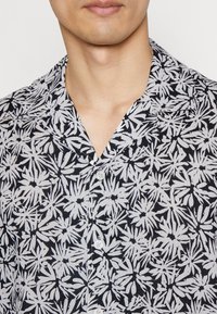 Hombre con una camisa de botones de patrón floral en blanco y negro, con el cuello abierto, mostrando el cuello y la parte inferior de la cara.