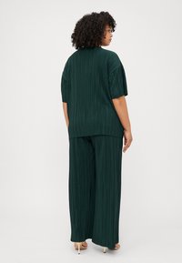 Άτομο με σγουρά μαλλιά σε ένα πράσινο πλισέ δύο κομματιών, γυρισμένο μακριά, δείχνοντας τη ροή και τον κομψό σχεδιασμό του ρούχου σε έναν απλό φόντο.