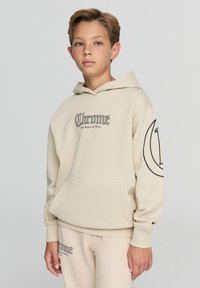 Beżowa bluza z kapturem z przednią kieszenią oraz czarnym tekstem graficznym "Chrome" i "The Ruler of Ways". Posiada ściągacze przy rękawach oraz kieszeń kangurzą.