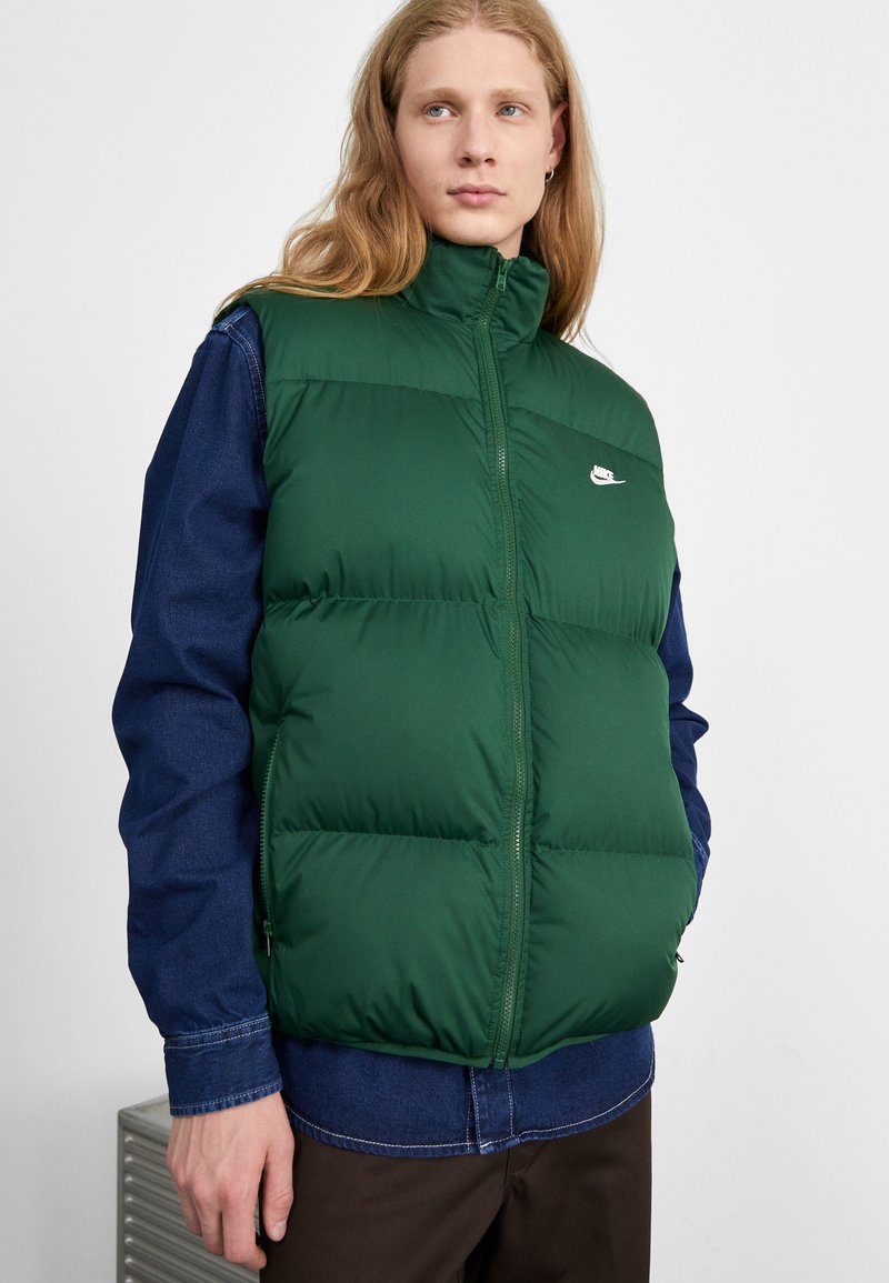 Grønne polstrede vest med høj krave, lynlåslukning, to sidelommer og et lille hvidt logo, båret over en blå denimskjorte.