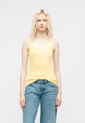 Kobieta o długich blond kręconych włosach, ubrana w żółtą, bezrękawnikową bluzkę na guziki i niebieskie dżinsy, stojąca na tle jednolitego jasnego tła.