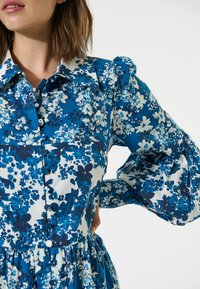 Ein Blumenkleid in Blau- und Weißtönen, mit Knopfleiste, großen Puffärmeln und einer gerafften Taille, aus einem glatten Stoff gefertigt.