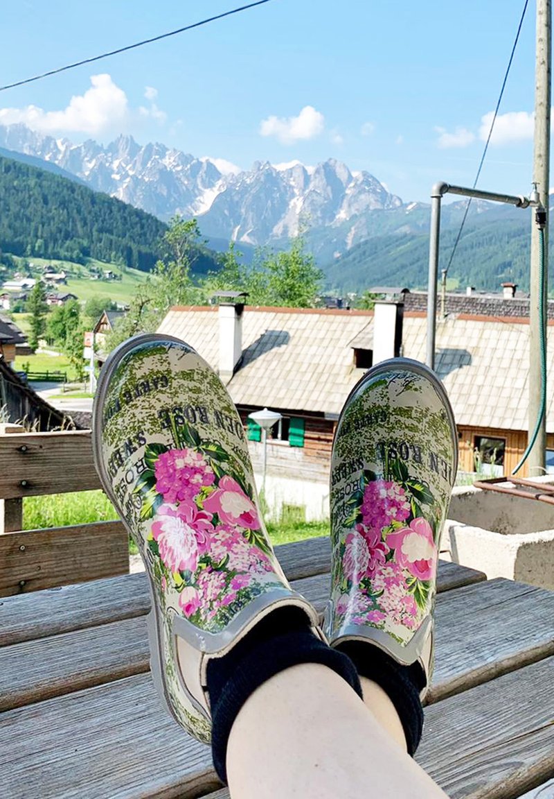Bottes de pluie en caoutchouc à motifs floraux avec des fleurs roses et violettes, sur fond vert et détails gris. Portées avec des chaussettes noires à la cheville, sur une surface en bois.