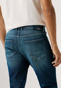 Vaqueros de mezclilla en azul oscuro con un corte ajustado. Incluyen cinco bolsillos, costuras en contraste y una etiqueta de cuero con la marca en la cintura.