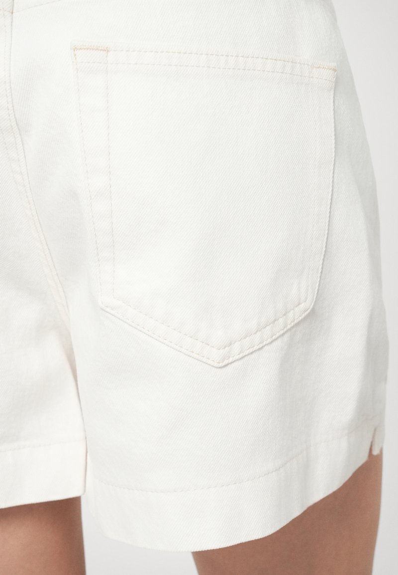 Shorts en denim blanc avec un revers replié, présentant une poche arrière unique avec des détails de couture visibles. La texture semble lisse et robuste.