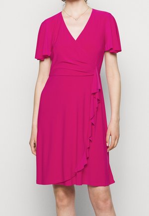 Mujer con vestido cruzado magenta hasta la rodilla, con mangas con movimiento y detalle de volantes a lo largo de la cintura y la falda.