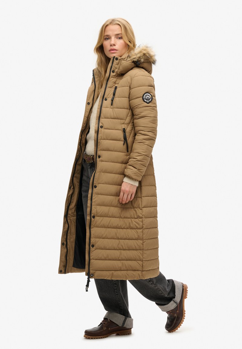 Langer, brauner Daunenmantel mit einer Kapuze aus Fellbesatz, Reißverschlusstaschen und einem gesteppten Design. Über einem cremfarbenen Pullover und dunklen Jeans getragen.