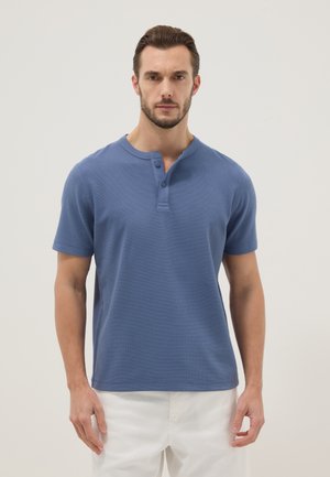Uomo con capelli castani corti e barba che indossa una maglietta henley blu a maniche corte e pantaloni bianchi, in piedi davanti a uno sfondo semplice.
