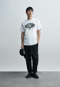 Λευκό κοντομάνικο μπλουζάκι με μαύρο λογότυπο "MOSCHINO", σε συνδυασμό με μαύρο παντελόνι με φερμουάρ και μαύρα αθλητικά παπούτσια. Κάσκα στο χέρι.