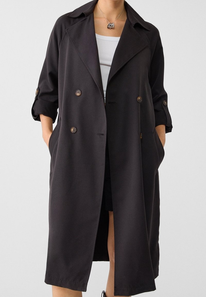 Femme portant un long manteau noir croisé avec les manches retroussées par-dessus un débardeur blanc et un collier, les mains dans les poches du manteau.