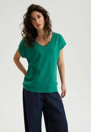 Kobieta o falowanych włosach, ubrana w zieloną bluzkę z krótkim rękawem i dekoltem w serek oraz luźne, ciemnoniebieskie spodnie, stojąca z jedną ręką na biodrze.