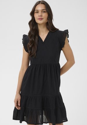 Žena s dlhými vlnitými vlasmi, oblečená v čiernych šatách bez rukávov s volánmi na ramenách a viacvrstvovou sukňou, stojaca pred jednoduchým pozadím.