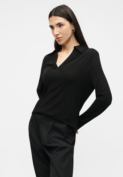 Γυναίκα με μακριά σκούρα μαλλιά, ντυμένη με μαύρο πουκάμισο μακριού μανικιού με γιακά και μαύρο παντελόνι, στέκεται με τα χέρια πίσω στην πλάτη σε λευκό φόντο.