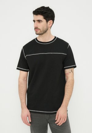 Άνδρας με σκούρα μαλλιά που φοράει μαύρο τζόκεϊ με λευκές ραφές και γκρι παντελόνι, κοιτάζοντας προς τα αριστερά μπροστά σε απλό φωτεινό φόντο.