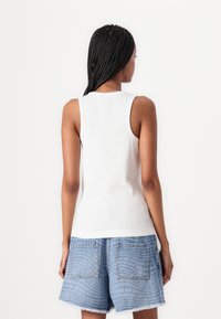 Hvit, ribbet tanktopp med rund halslinning og brede skulderstropper, sammen med blå denimshorts med et trykt mønster.