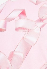Vaaleanpunainen kangas, jossa on kiiltävä pinta, esittelee suuria satiinisetäkinauhoja eri kokoisin, jotka on kehystetty hienovaraisilla ompeluilla.