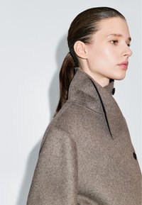 Cappotto di lana marrone con colletto alto, caratterizzato da una chiusura a pulsante singolo e una trama morbida, esposto su uno sfondo chiaro.