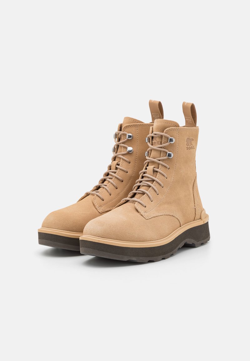 Sandfarbene Wildleder-Ankle-Boots mit abgerundetem Zehenbereich, Metallösen und einer dicken, dunklen Gummisohle. Mit einer kleinen Zuglasche auf der Rückseite.