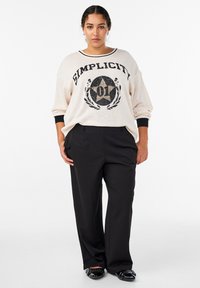 Krēmkrāsas oversize džemperis ar melnu tekstu un zvaigznes grafiku, kopā ar melnām pieguļošām biksēm, valkāts ar spīdīgiem melniem loaferiem.