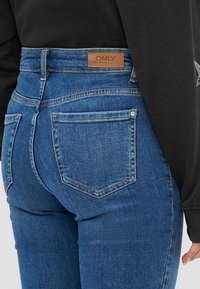 Nærbilde av en person som har på seg blå høytlivs jeans med baklommer og et brunt lærmerkelapp, kombinert med en svart langermet topp.