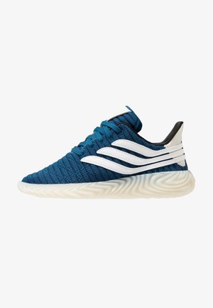 Zapatilla deportiva de tela azul con acentos ondulados blancos, parte superior texturizada y una suela de goma blanca flexible con un patrón en zigzag.