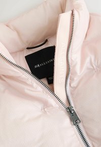 Lichtroze pufferjack met een gewatteerde textuur, zilveren rits en een label met "ALLSAINTS." De binnenvoering is soepel en zacht.