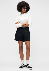 Femme aux cheveux afro portant des lunettes de soleil noires, un haut blanc à manches longues, un short noir, des chaussettes blanches et des baskets noires, debout les bras croisés.