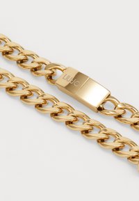 Zlatý náramok na reťaz s hrubými článkami a obdĺžnikovou sponou s vygravírovaným logom "LIU•JO" na bielom pozadí.