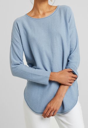 Vrouw draagt een lichtblauwe trui met lange mouwen en een ronde hals, met een witte broek, armen gekruist ter hoogte van de taille.