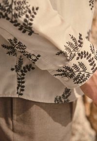 Světle barevná košile s černou výšivkou kapradí, s manžetami na knoflíky a jemnou texturou, doplněná neutrálně zbarvenými kalhotami.