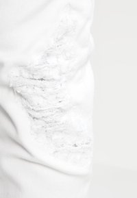 Białe dżinsy zniszczone na udzie, z postrzępioną łatą, podkreślającą kontrast faktur z gładkim materiałem.