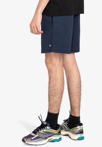 Marina shorts i mjukt tyg, med en sidficka och en liten logotyp vid fållen. Matchade med flerfärgade löparskor och svarta strumpor.