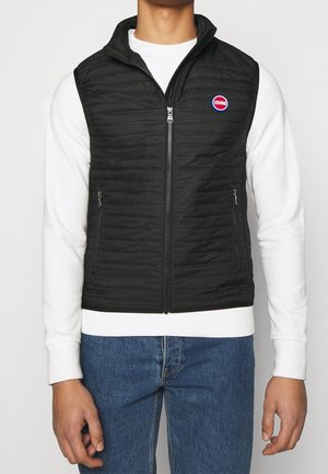 Mężczyzna ubrany w czarną pikowaną kamizelkę na zamek, na białej koszuli z długim rękawem i niebieskich dżinsach, stojący na jednolitym tle.