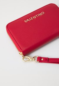 Raudona odinė piniginė su auksiniu "Valentino" logotipu, užtrauktuko uždarymas ir nuimama dirželis. Lygus paviršius ir plonas stačiakampis dydis.